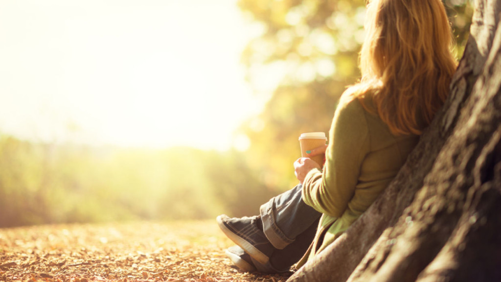 Woman with reddish hair sitting against a tree trunk holding a coffee cup in warm golden hour sunlight.