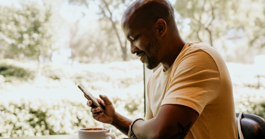 Man in yellow t-shirt looking down at his phone with blurred green foliage in the background during daytime.