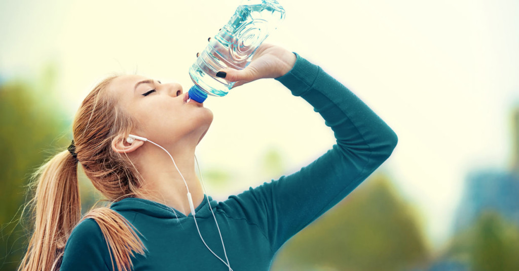 Woman with reddish hair in a ponytail wearing a teal athletic jacket and earbuds, drinking from a water bottle outdoors with blurred green background