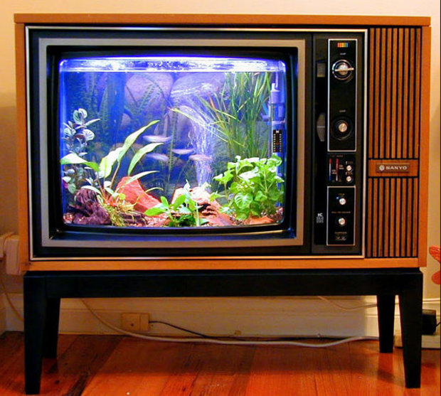 Vintage wood-paneled Sanyo television console converted into an aquarium, displaying colorful fish, plants, and coral inside the screen, mounted on a black stand.