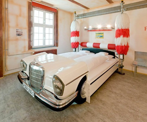 Bedroom with a bed shaped like a vintage white Mercedes-Benz car, featuring red and white striped fabric canopy above, wooden flooring, and a window with red frame.