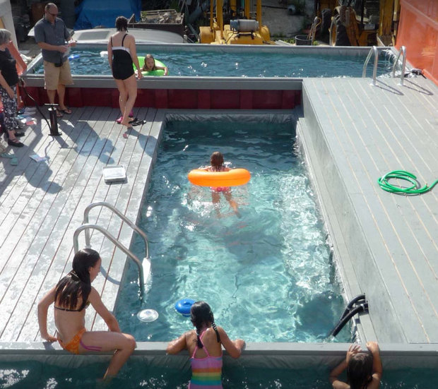 Overhead view of children and adults at a swimming pool, with a child floating on an orange ring in clear blue water and others sitting on the wooden deck.