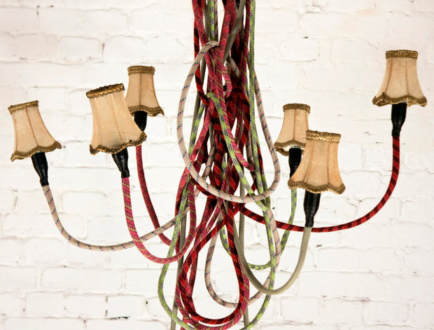 Handmade chandelier made from colorful braided rope in red, green, and cream with small fabric lampshades attached to the ends, hanging against a white brick wall.