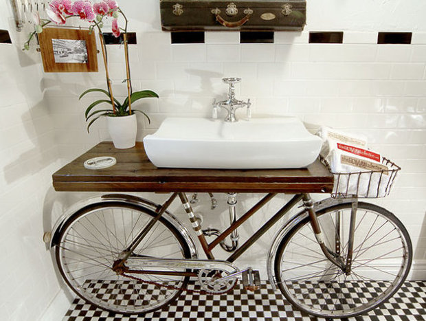 Bathroom sink made from a repurposed bicycle frame with a wooden shelf holding a white rectangular basin, potted orchid plant, and checkered floor.