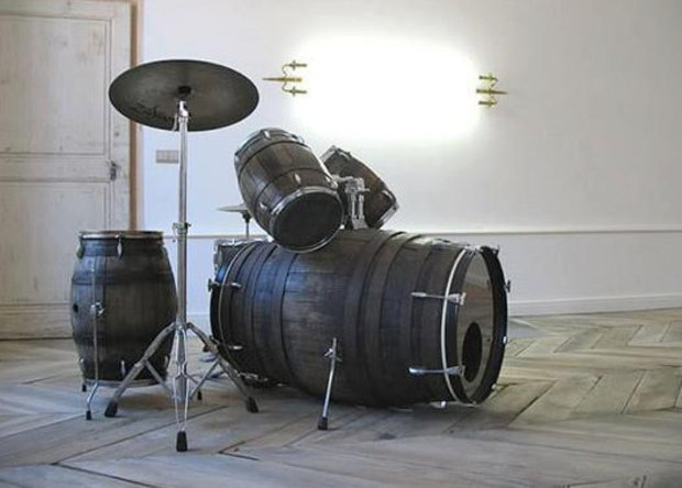 Drum kit made from black recycled plastic barrels and oil drums with metal bands, displayed in a white gallery space with wooden flooring.
