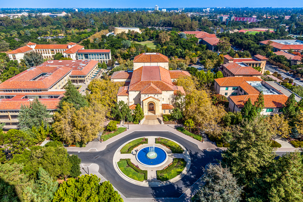 Aerial view of stanford university campus with a domed central building, circular plaza with blue fountain, surrounded by green trees and orange-roofed buildings.
