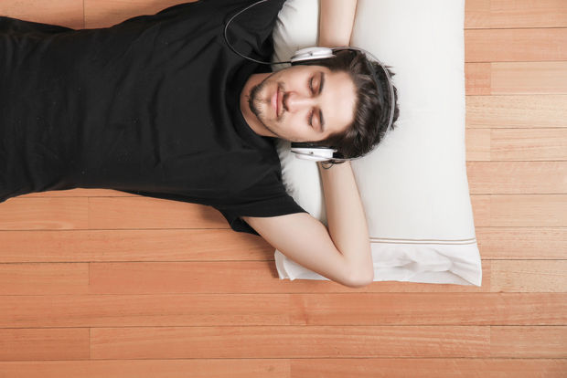 Person lying on a wooden floor wearing headphones around their neck, relaxing and smiling while resting on a white pillow.