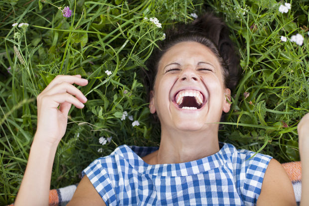 Person laughing while lying on their back in grass and wildflowers, wearing a blue and white checkered shirt.