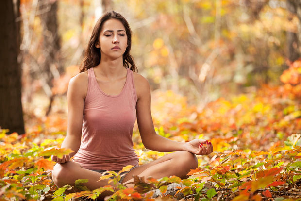 Meditation Woman in pink tank top sitting in meditation pose surrounded by orange and yellow fall leaves in a forest.