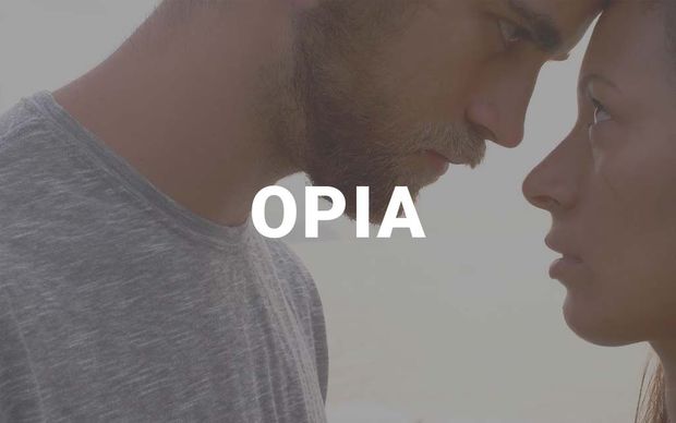 A close-up profile shot of a man and woman facing each other intimately, foreheads nearly touching, with the word 'OPIA' displayed in white capital letters in the center — a soft, muted-tone image.