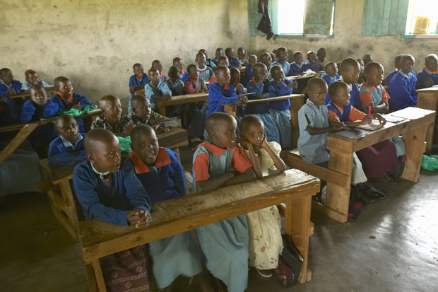 Large classroom full of African students wearing blue uniforms seated at wooden desks in a basic school building.