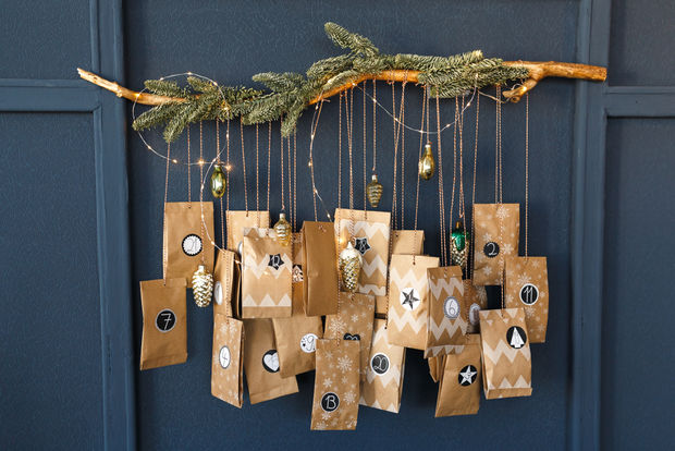 Handmade Advent calendar with numbered kraft paper bags hanging from a natural branch with gold ornaments and decorative elements against a dark blue wall.