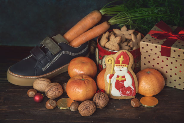Child's shoe surrounded by traditional Sinterklaas items including carrots, walnuts, oranges, cookies, and a Saint Nicholas chocolate figure with gift boxes.