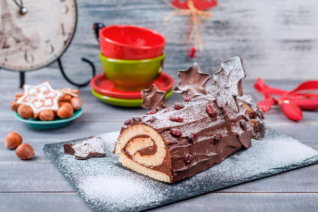 Chocolate Yule log cake dusted with powdered sugar decorated with chocolate leaves, surrounded by small cookies, colorful bowls, and festive holiday decorations on a wooden surface.