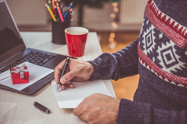 Person in a festive Nordic-patterned sweater writing on a notepad at a desk with a laptop, red mug, and small gift box in warm holiday lighting.