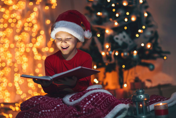 Child in a Santa hat and red shirt reading a book surrounded by Christmas tree lights with warm golden bokeh background.