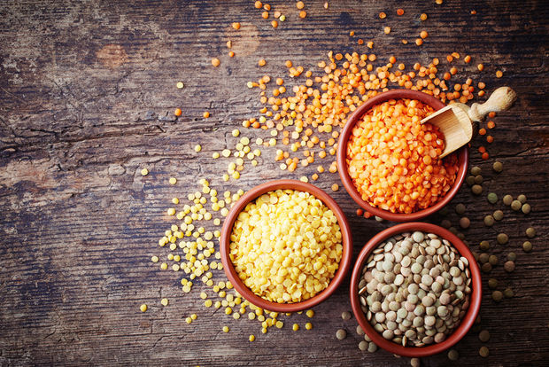 Three terracotta bowls containing different varieties of lentils — red lentils, yellow split lentils, and green/brown lentils — arranged on a dark rustic wooden surface with a wooden scoop and scattered lentils.
