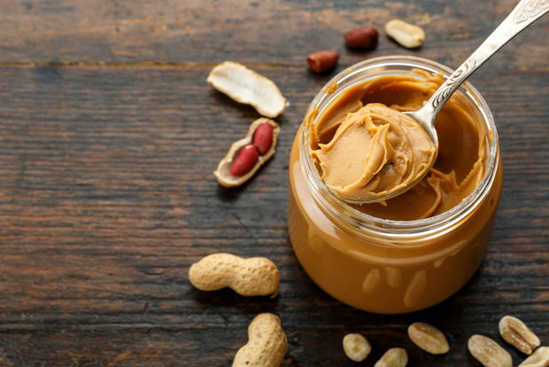 An open glass jar of creamy peanut butter with a silver spoon scooping out a dollop, surrounded by whole and shelled peanuts on a dark wooden surface