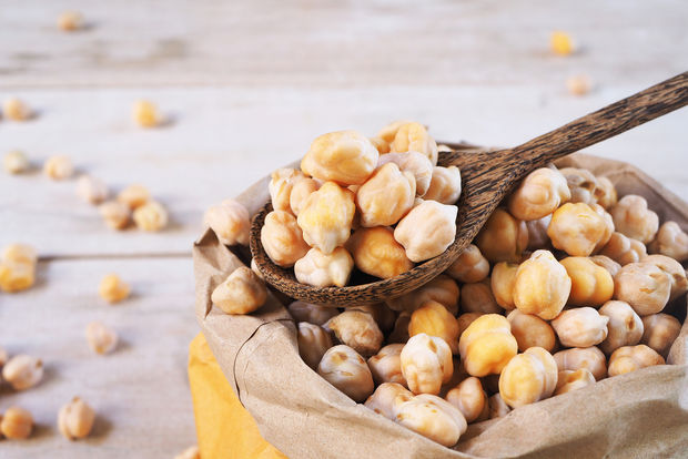 Dried chickpeas spilling from a brown paper bag with a wooden spoon scooping them up, on a light wooden surface with scattered chickpeas in the background.