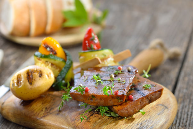 Grilled marinated seitan steaks garnished with fresh thyme and pink peppercorns, served on a round wooden board with a colorful vegetable skewer and grilled onions, with sliced bread in the background.