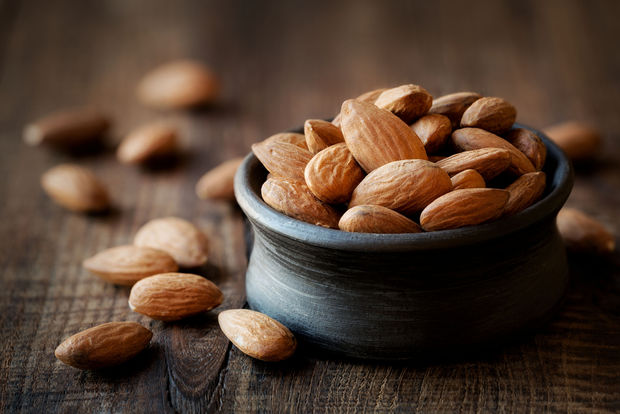 Raw whole almonds in a small dark blue ceramic bowl on a rustic wooden surface, with several almonds scattered beside the bowl.