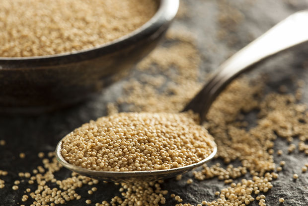 A silver spoon overflowing with tiny golden amaranth seeds in the foreground, with a rustic metal bowl filled with more amaranth in the background, on a dark slate surface with scattered seeds.