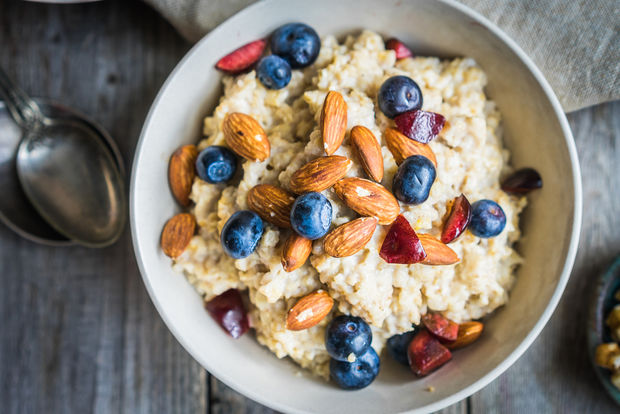 A bowl of creamy oatmeal topped with fresh blueberries, whole almonds, and sliced strawberries, viewed from above on a rustic wooden table with a silver spoon and linen napkin beside it.
