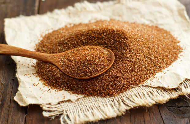 A pile of reddish-brown teff grain spread on a cream-colored linen cloth, with a wooden spoon resting in the center, on a dark rustic wooden table.