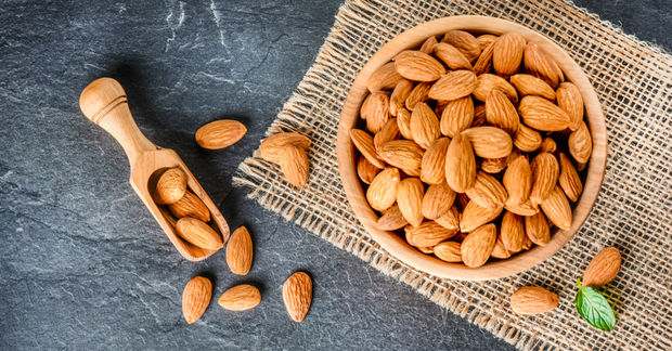 A wooden bowl filled with whole almonds and a small wooden scoop on burlap cloth over a dark slate surface.