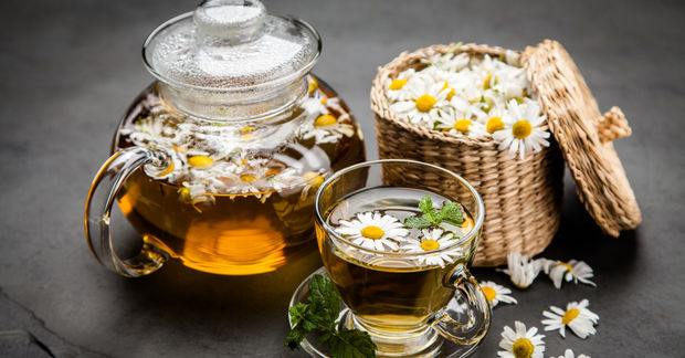 A glass teapot and cup of chamomile tea with floating daisies, beside a woven basket of fresh chamomile flowers.
