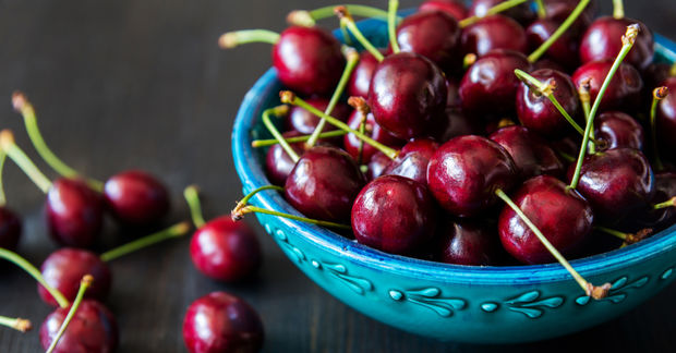 Fresh dark red cherries with green stems overflowing from a turquoise ceramic bowl on a dark wooden surface.