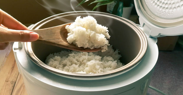 A hand lifts a wooden spoon of steaming white rice from an open rice cooker, with steam rising from the pot.