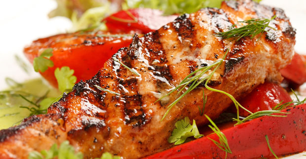A grilled salmon fillet with charred grill marks, garnished with fresh dill and herbs, served with red peppers and greens.