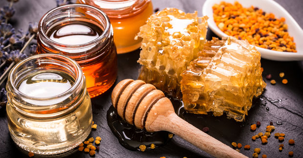 Honeycomb, jars of different honey varieties, a wooden dipper, and bee pollen on a dark slate surface.