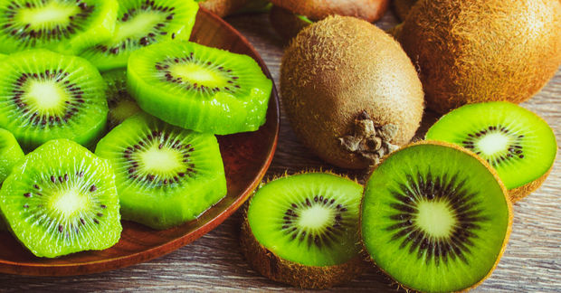 Sliced green kiwi fruit on a wooden plate alongside whole and halved kiwis on a light wooden surface.