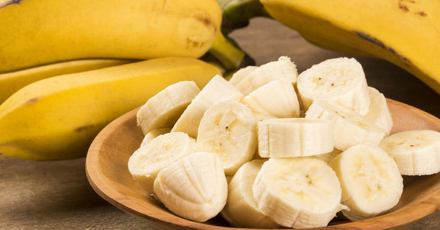 Sliced banana pieces on a wooden plate with whole ripe yellow bananas in the background on a wooden surface.