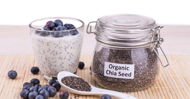 A glass jar labeled 'Organic Chia Seed' with a metal clasp lid, alongside a glass of chia seed pudding topped with fresh blueberries, a white ceramic spoon filled with chia seeds, and scattered blueberries on a bamboo mat.