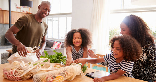 A happy family unpacks fresh groceries from reusable bags onto a kitchen table in a bright living space.