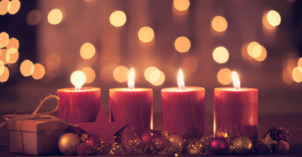 Four lit red candles with warm golden bokeh lights in the background, surrounded by holiday ornaments, greenery, and a small gift box.