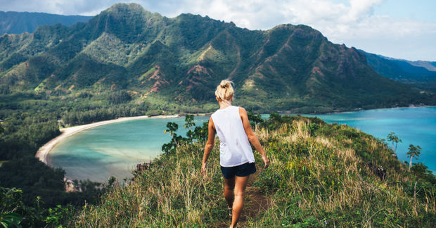 photo Woman in white walking on mountain trail overlooking tropical coastline with turquoise water and lush green landscape.