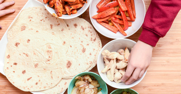 Child's hand reaching for cashews while assembling a wrap or taco with tortilla, cooked bacon, red bell peppers, and other ingredients on a wooden surface.