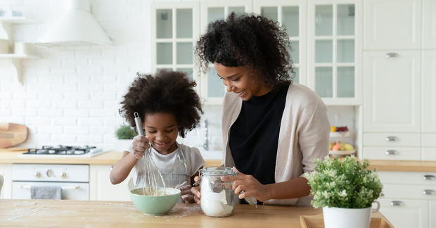 Woman and child baking together in a bright white kitchen, with the child wearing an apron and stirring a bowl of ingredients.