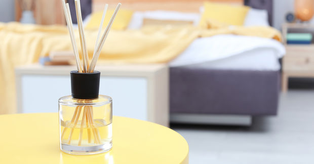 A reed diffuser with wooden sticks in a glass bottle sits on a yellow table in a cozy bedroom.