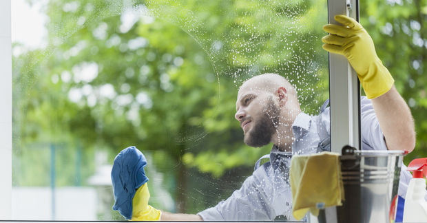 A man wearing yellow rubber gloves washes a large window with a blue cloth and soapy water.