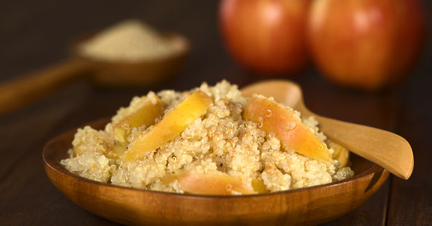 Bowl of quinoa topped with dried apricot slices with a wooden spoon, with blurred onions in the background.