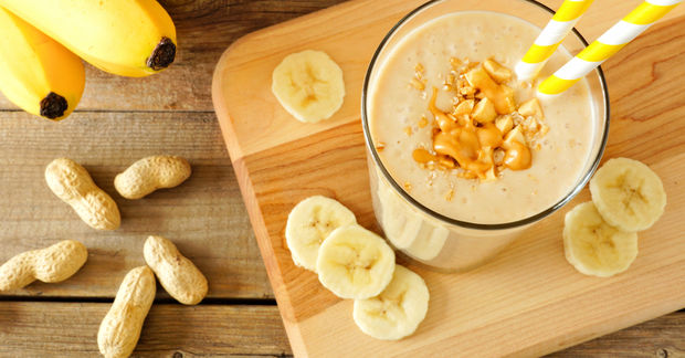 Banana peanut butter smoothie topped with granola in a glass, surrounded by banana slices, whole peanuts, and fresh bananas on a wooden board.