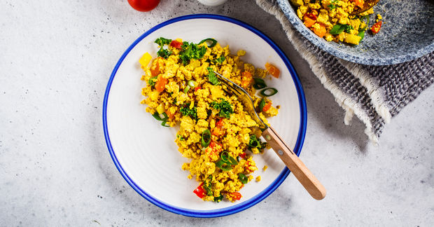 Scrambled eggs with corn, red peppers, and green herbs in a white bowl with blue rim on a concrete surface.