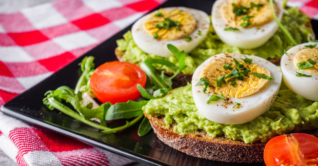 Halved hard-boiled eggs topped with herb filling on avocado-spread bread slices with tomato garnish on a black plate with checkered cloth.