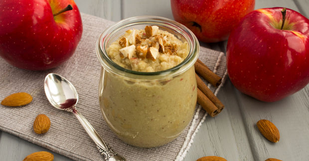 Glass jar of apple cinnamon yogurt topped with almond slices, surrounded by fresh red apples, almonds, cinnamon sticks, and a spoon on a white cloth.