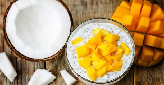 Bowl of coconut chia seed pudding topped with fresh mango chunks, with a coconut half and fresh mango visible alongside on a wooden surface.